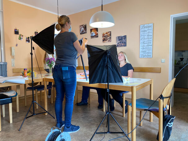 Zwei Personen sitzen am Tisch und eine Frau beleuchtet sie.